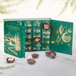 A green Gearharts Fine Chocolates Christmas advent calendar with gold illustrations, shown open with several chocolates placed in front on a light marble surface.