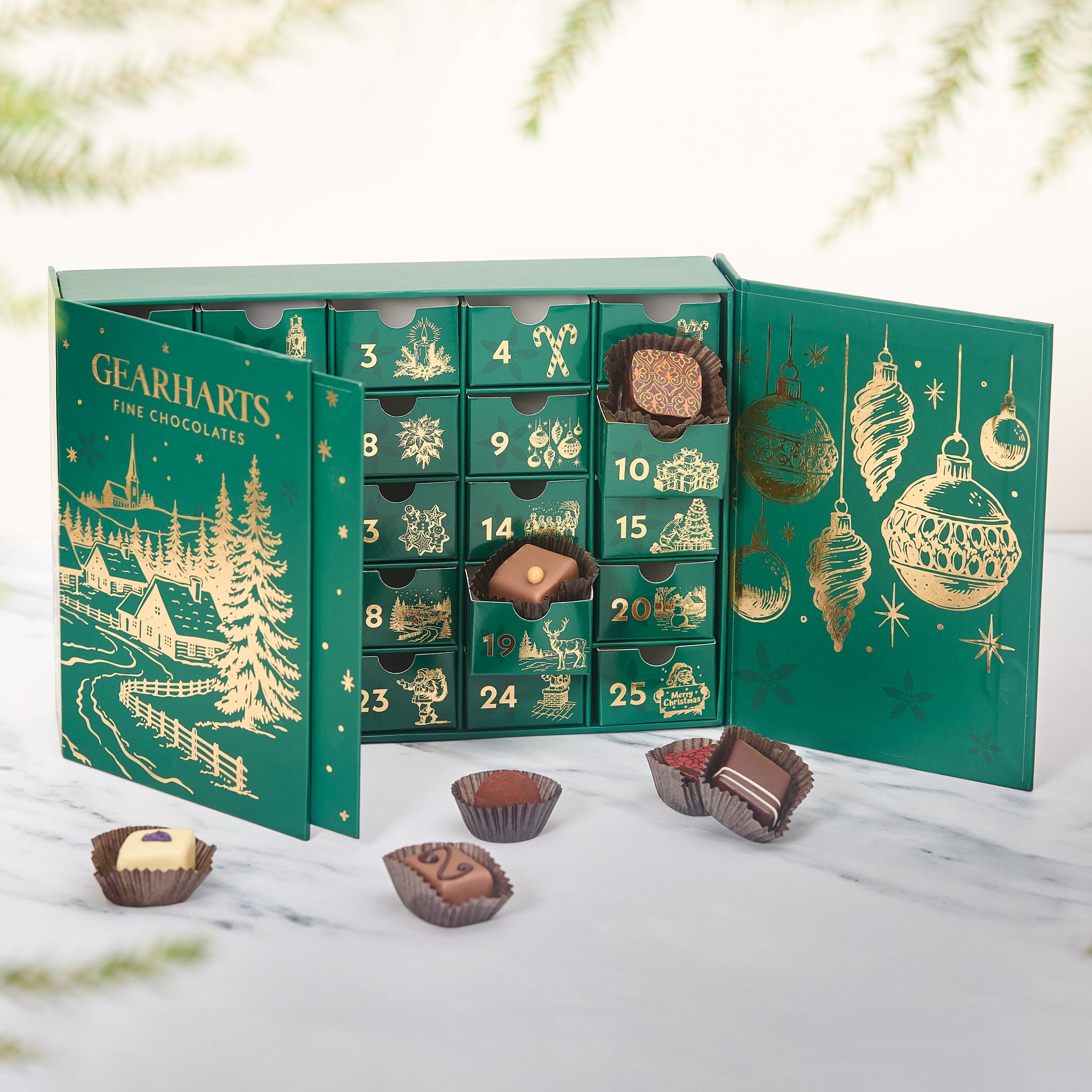 A green Gearharts Fine Chocolates Christmas advent calendar with gold illustrations, shown open with several chocolates placed in front on a light marble surface.
