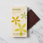 A boxed ginger dark chocolate bar from Gearharts Fine Chocolates sits on a light marble surface next to a partially unwrapped dark chocolate bar. The box is cream-colored with green and yellow floral illustrations and reads 'GINGER' and 'GEARHARTS FINE CHOCOLATES.