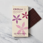 A box of Gearharts Criolla dark chocolate bar with pink and purple floral graphics, shown next to a partially unwrapped dark chocolate bar on a white marble surface.