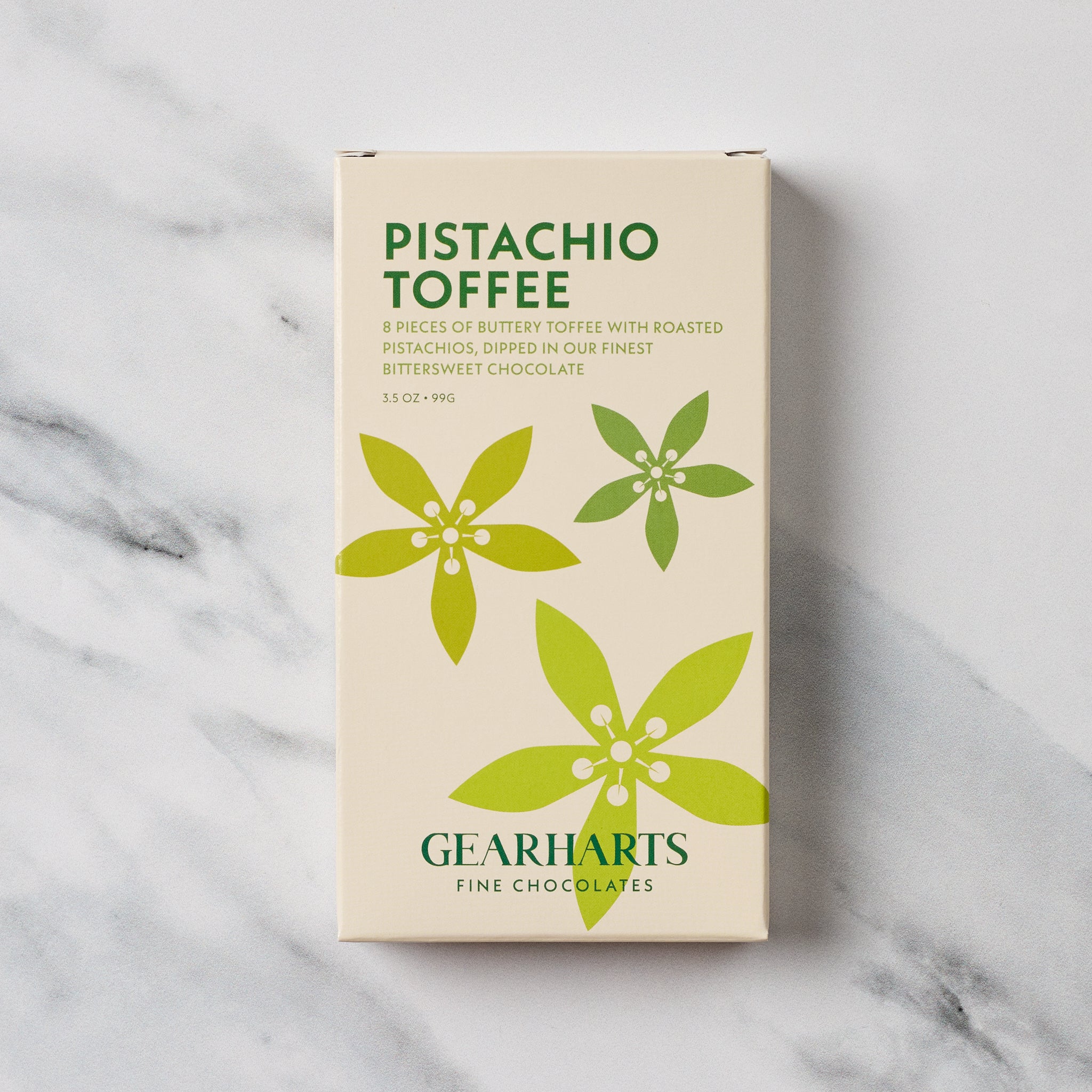 A boxed bar of Gearharts Pistachio Toffee chocolates, featuring a cream-colored box with green text and floral graphics, set against a light gray marble background.