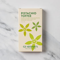 A boxed bar of Gearharts Pistachio Toffee chocolates, featuring a cream-colored box with green text and floral graphics, set against a light gray marble background.