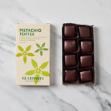 A box of Gearharts Pistachio Toffee chocolates next to an open tray of twelve individually wrapped dark chocolate toffees on a white marble surface.