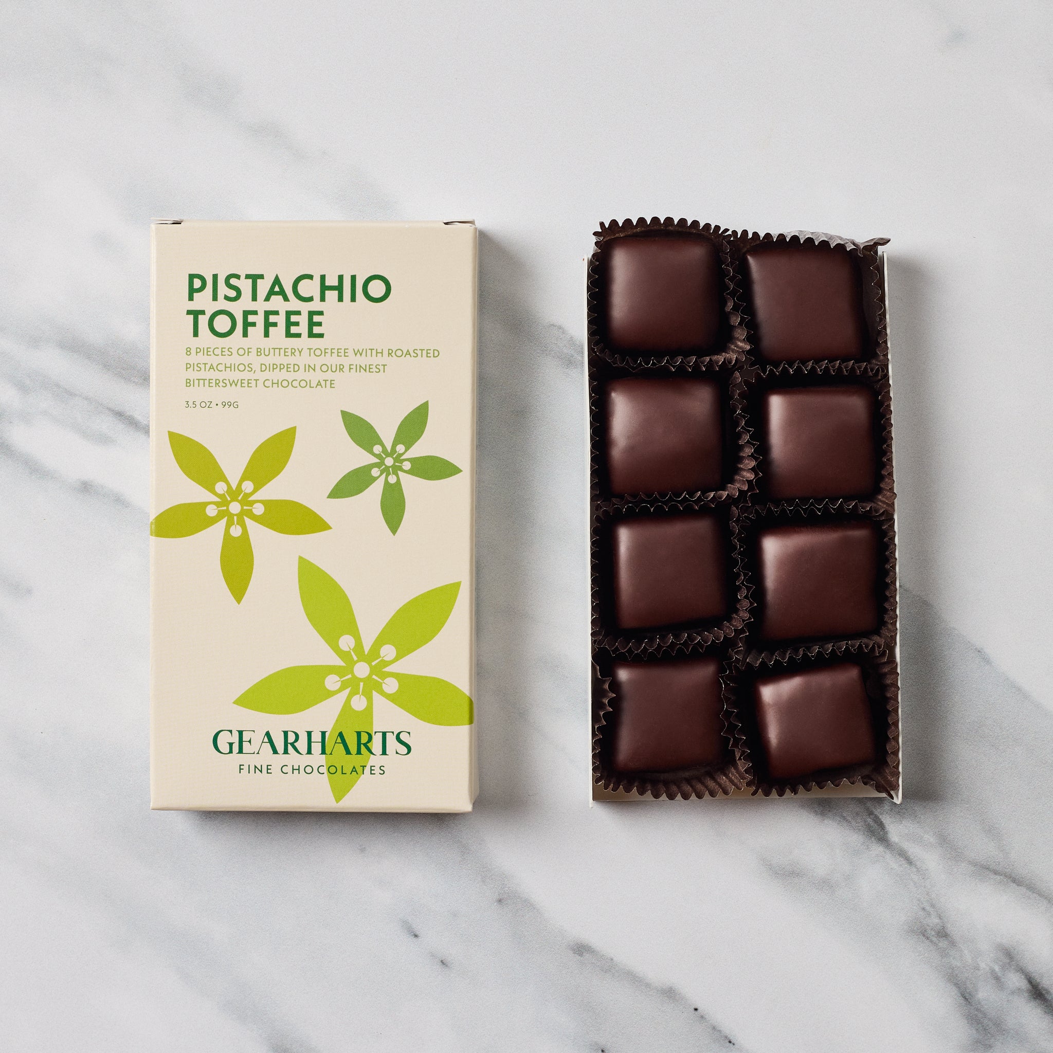 A box of Gearharts Pistachio Toffee chocolates next to an open tray of twelve individually wrapped dark chocolate toffees on a white marble surface.