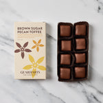 A box of Gearharts Brown Sugar Pecan Toffee alongside an open tray of individually wrapped chocolate toffees on a white marble surface.