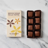 A box of Gearharts Brown Sugar Pecan Toffee alongside an open tray of individually wrapped chocolate toffees on a white marble surface.