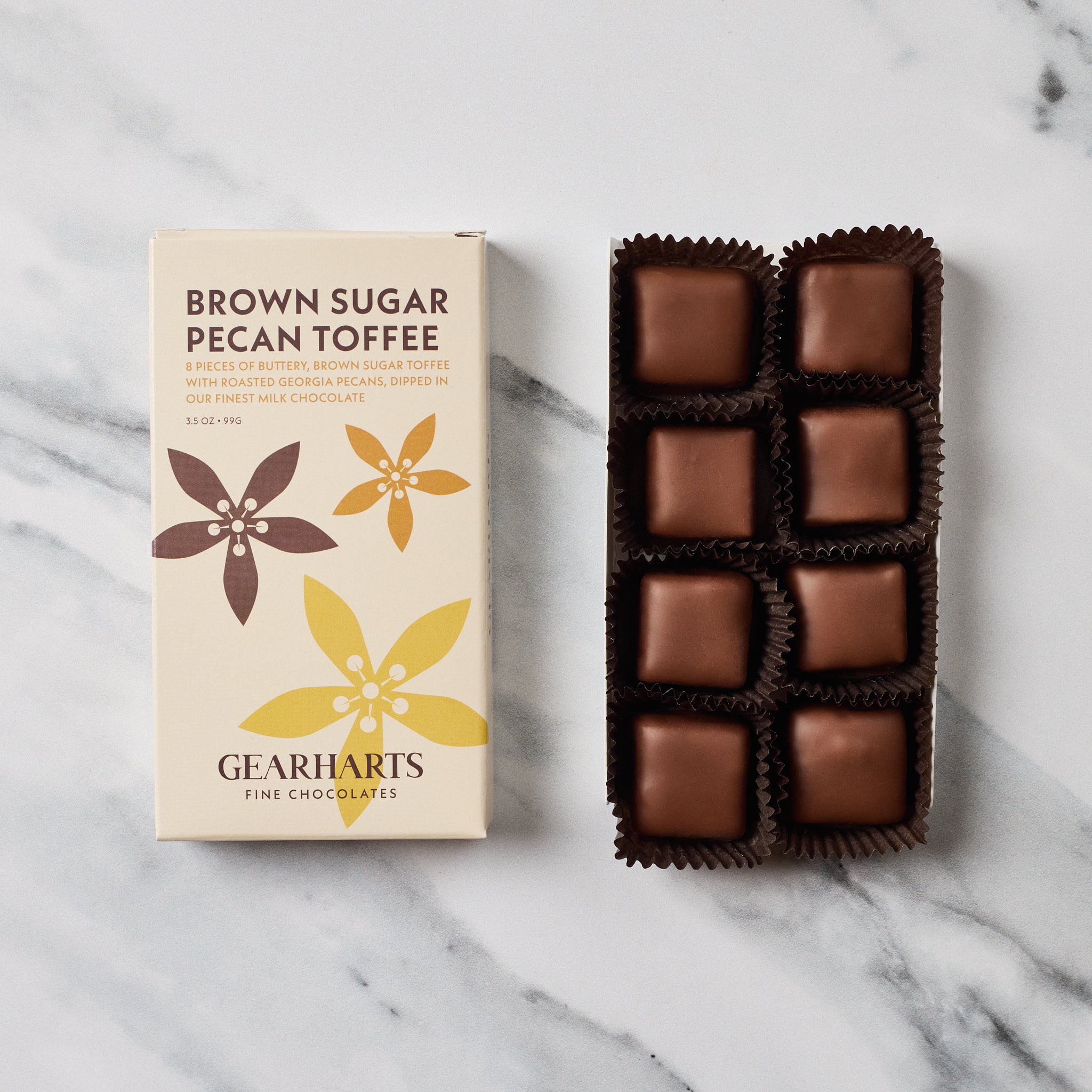 A box of Gearharts Brown Sugar Pecan Toffee alongside an open tray of individually wrapped chocolate toffees on a white marble surface.