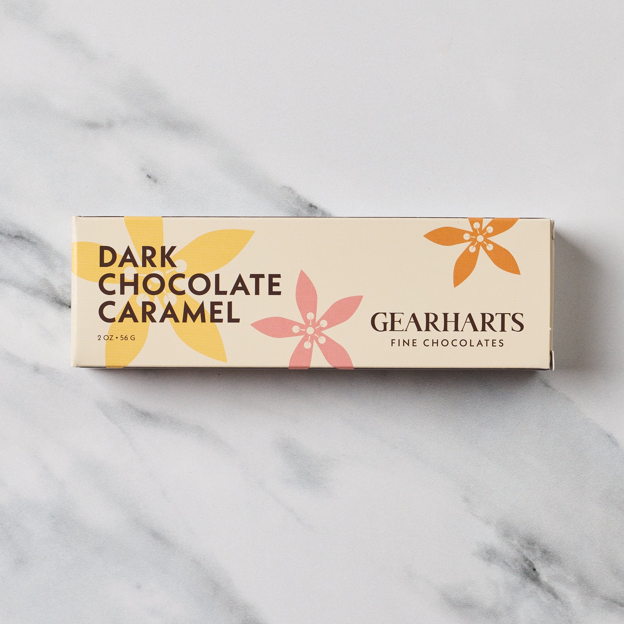 A rectangular box of Gearharts Fine Chocolates Dark Chocolate Caramel, featuring a cream-colored background with yellow, pink, and orange floral illustrations, set on a white marble surface.