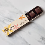 An open box of Gearharts Fine Chocolates Dark Chocolate Caramel truffles on a white and gray marble surface. The box is cream-colored with yellow and pink floral accents, and three chocolate truffles are visible inside.