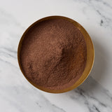 A top-down view of a fine brown powder, likely cocoa powder or coffee grounds, piled in a round gold-colored metal bowl on a white marble surface with gray veining.