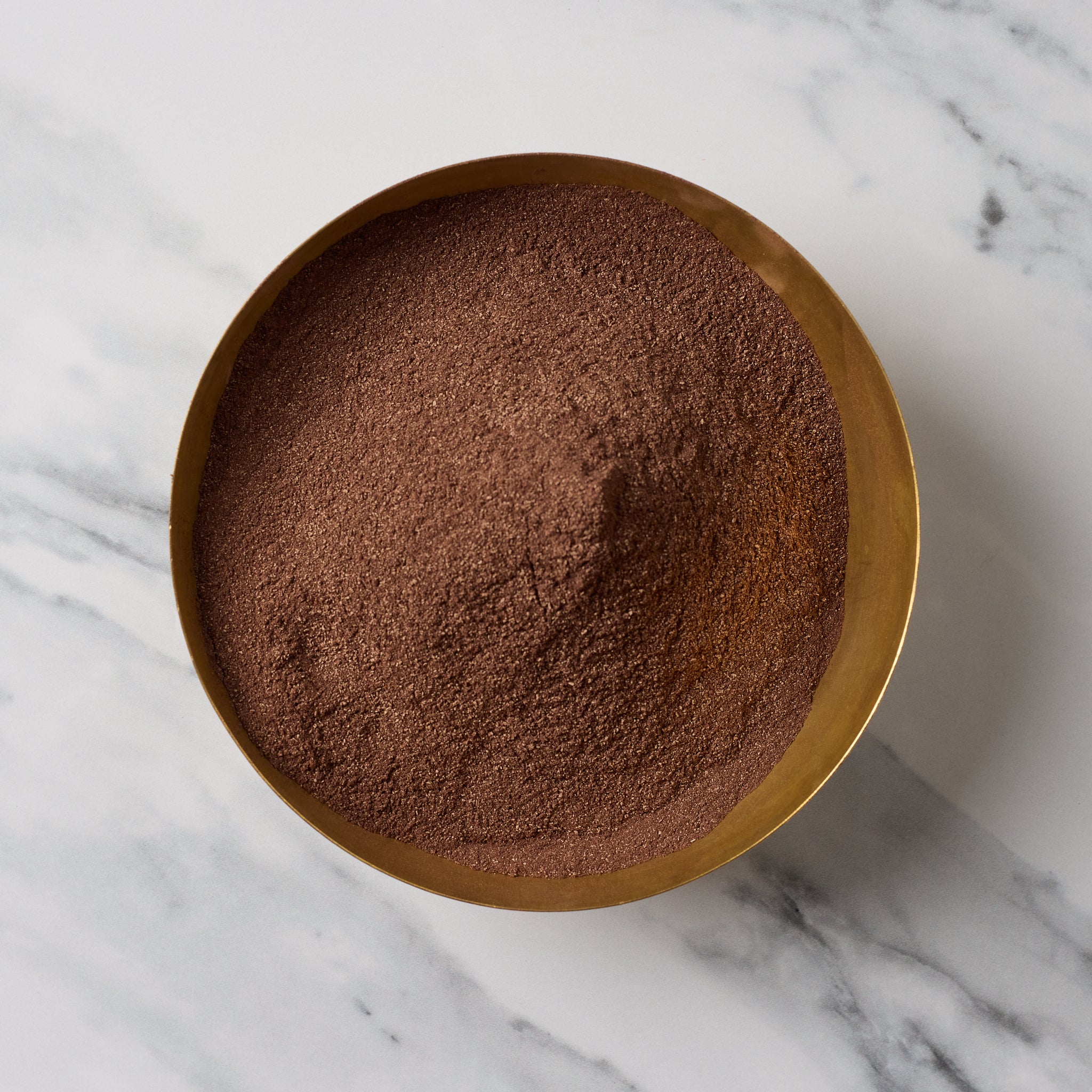 A top-down view of a fine brown powder, likely cocoa powder or coffee grounds, piled in a round gold-colored metal bowl on a white marble surface with gray veining.