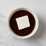 A white ceramic mug filled with a dark brown hot chocolate or cocoa drink, topped with a single white marshmallow, photographed from above on a light marble-like surface.