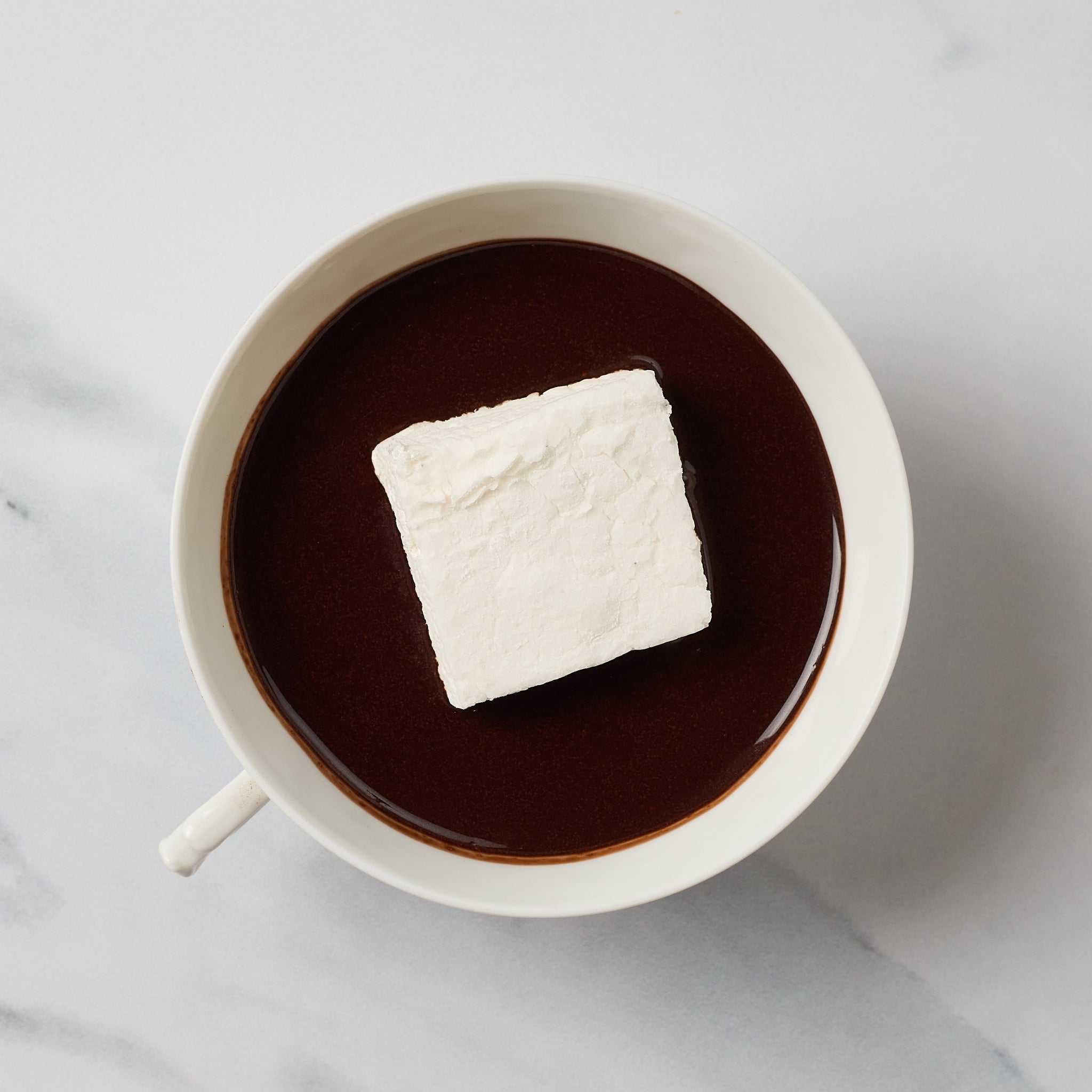 A white ceramic mug filled with a dark brown hot chocolate or cocoa drink, topped with a single white marshmallow, photographed from above on a light marble-like surface.