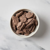 Chocolate-coated pecans in a small off-white ceramic bowl, photographed from above on a light marble surface.