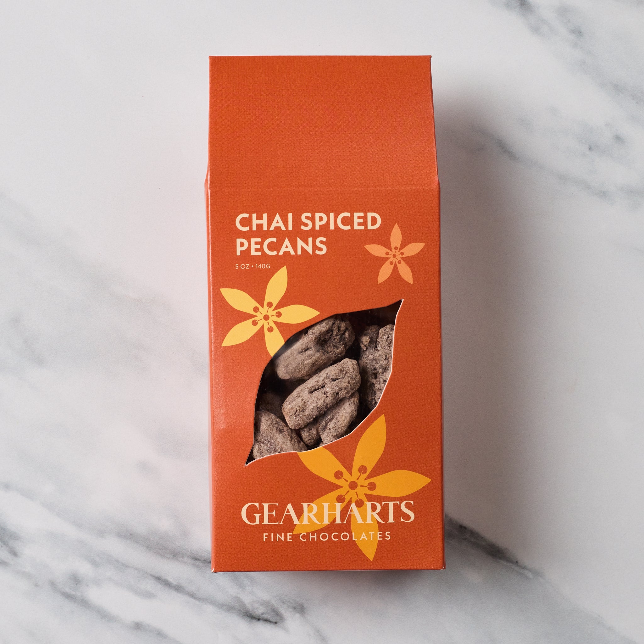 A rectangular orange box of Gearharts Chai Spiced Pecans with a clear window showing the pecans inside, set on a light gray marble surface.