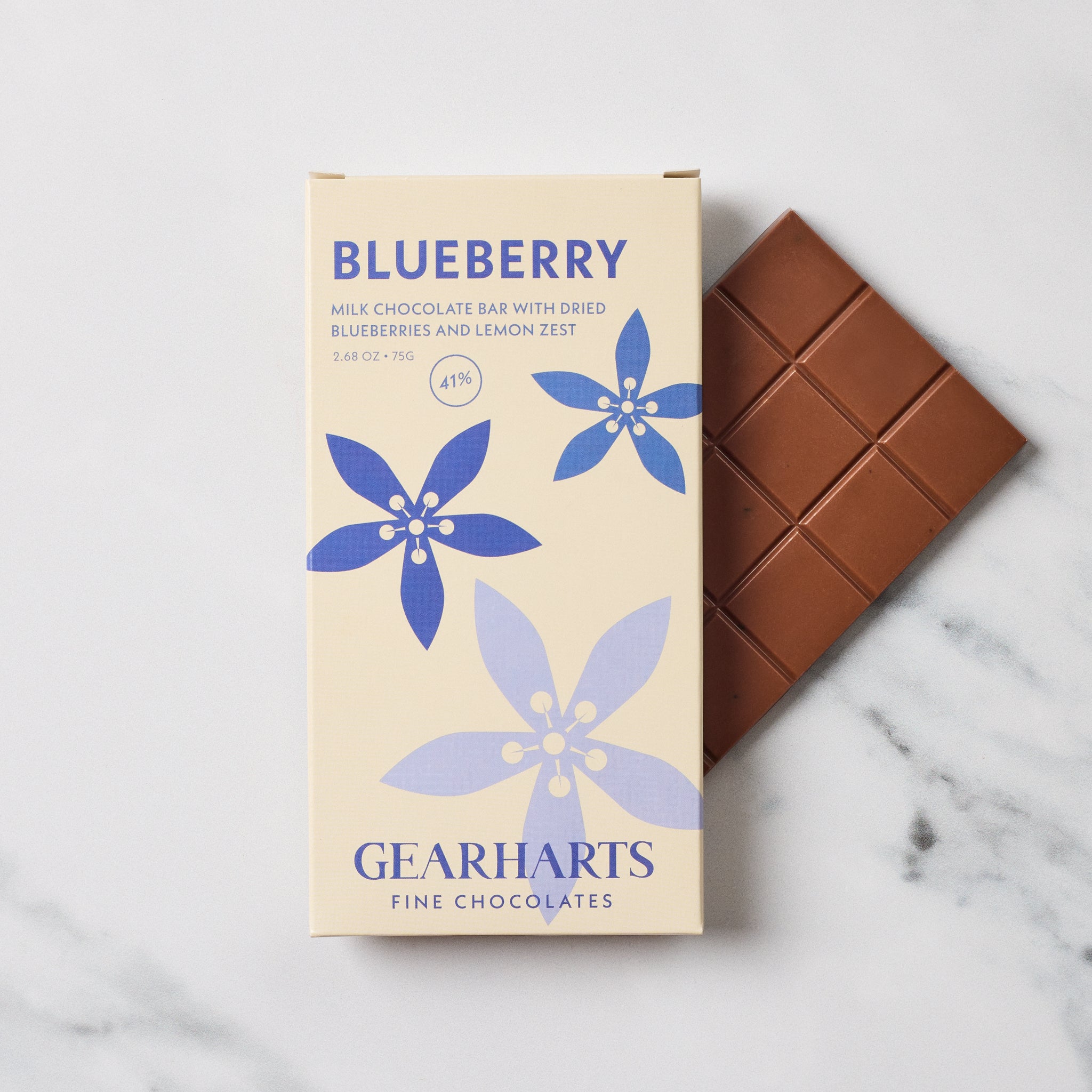 A box of Gearharts Blueberry milk chocolate bar with dried blueberries and lemon zest, shown next to a partially unwrapped milk chocolate bar on a light marble surface.